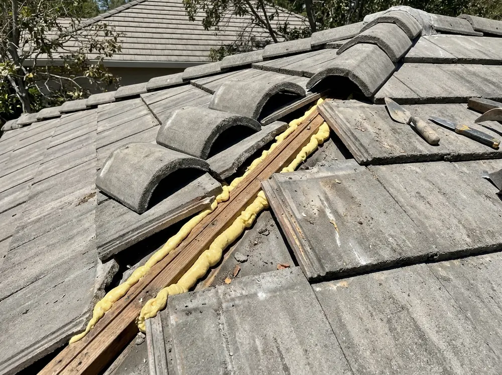 Hip caps lifted off a concrete tile roof showing beads of yellow foam adhesive along a ridge board